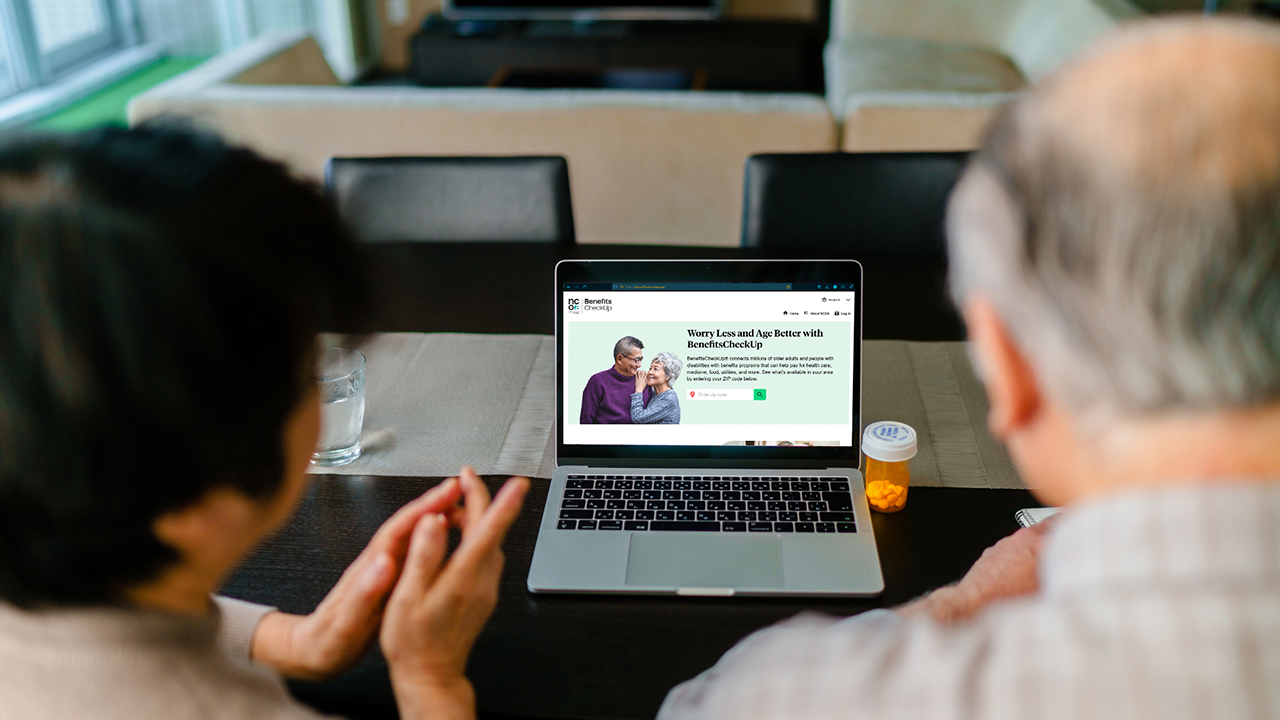 An older couple sits at a dining table, viewing a website on a laptop. The screen displays an NCOA BenefitsCheckUp page with the headline 'Worry Less and Age Better with BenefitsCheckUp.' The man has gray hair and wears a light-colored shirt, while the woman, partially obscured, has dark hair. On the table next to the laptop is a prescription bottle and a glass of water. The background features a living room with a sofa and a television.