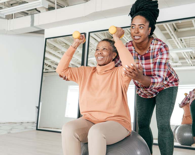 Black woman sitting on exercise ball in gym with hand weights and physical therapist helping her