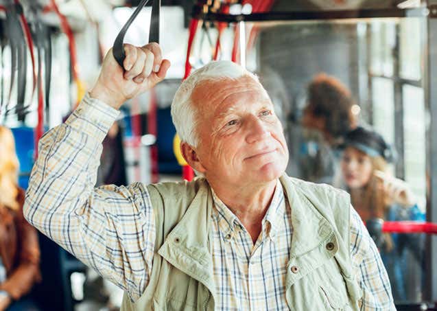 A senior man, wearing plaid and other outerwear, is standing up on a bus and enjoying his day.