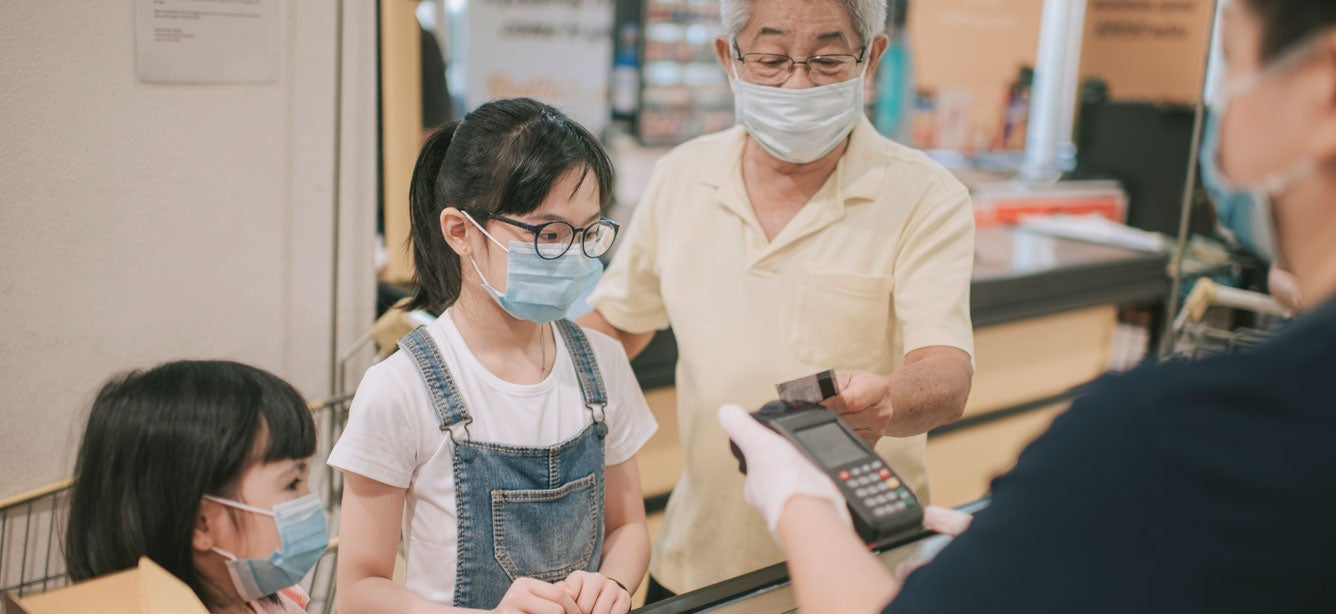 Senior Asian man is paying at the grocery register with his two granddaughters, while all are wearing their masks during the pandemic.