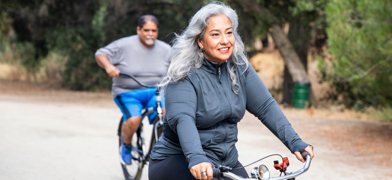 A senior Hispanic couple is outside biking together in nature.