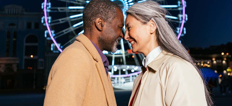 A senior biracial couple is standing in front of a ferris wheel at night with their foreheads touching.