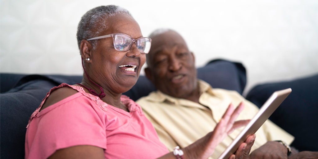 A Black senior woman and her husband are sitting on the couch looking at something on their tablet.
