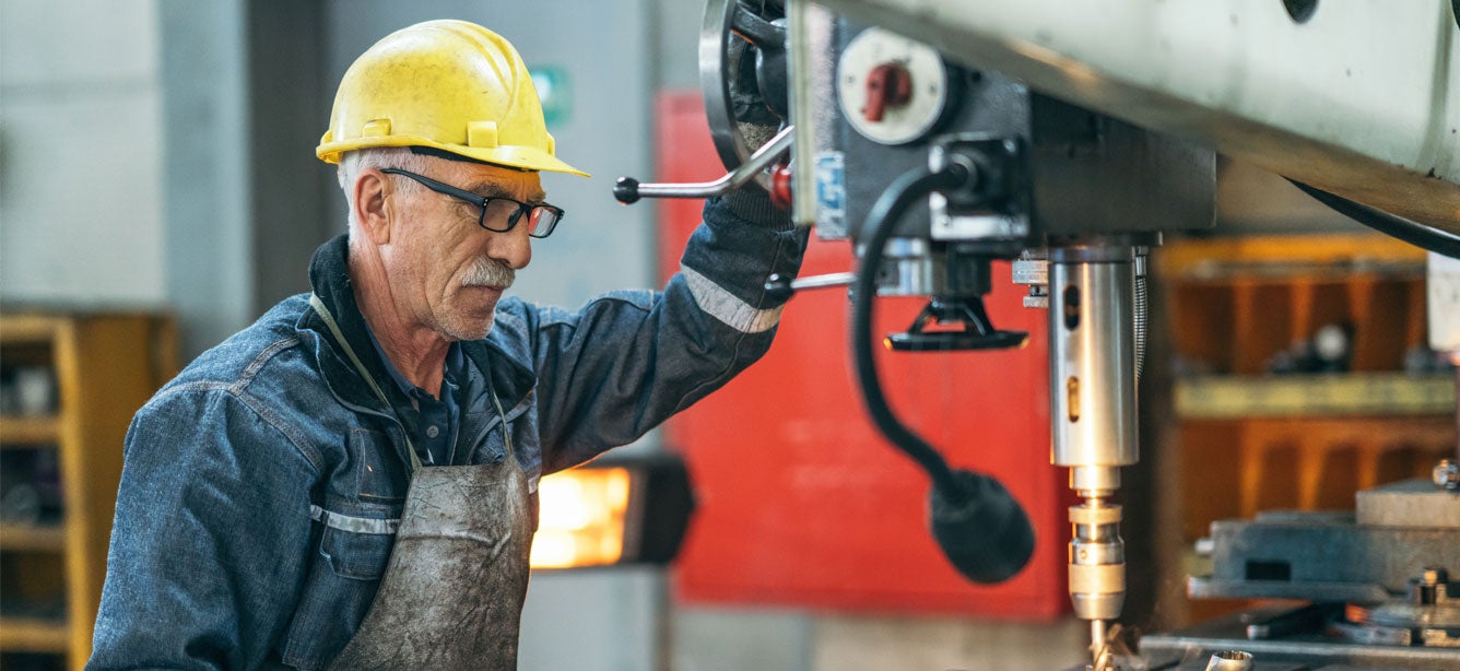 A senior man with a yellow hard hat and glasses works with a large drill bit at a plant workshop.