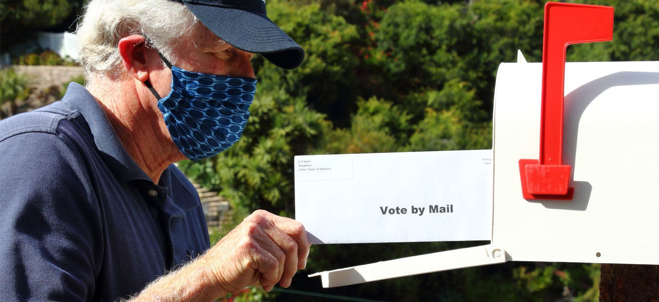 An older masked man is placing his ballot for the 2020 election in the mailbox.
