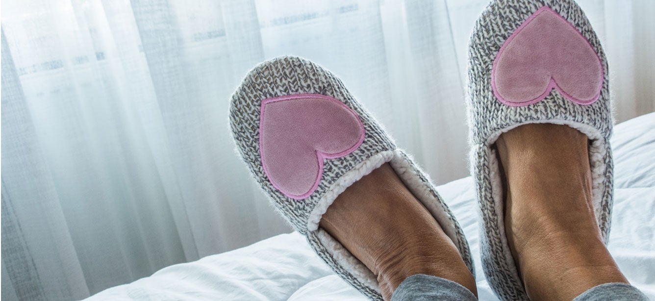 A senior woman's feet are pictured on a bed with her slippers on, where the slippers have pink hearts.