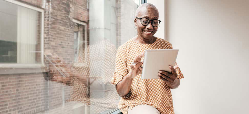 A senior Black woman wearing glasses rests near her window, using her tablet.