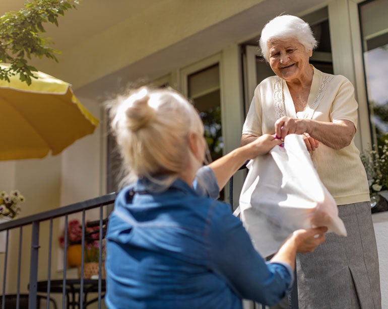 A middle-aged female Caucasian caregiver is handing groceries to her older mother while she's on the porch.