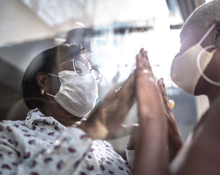 A Black female caregiver lovingly places her hands on the glass while her senior loved one is on the other side of the glass. 