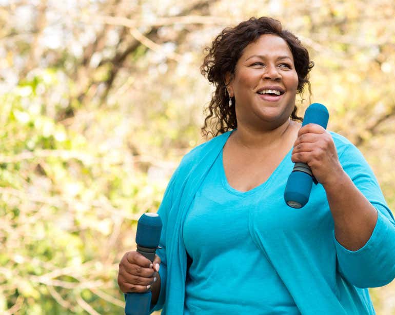 A senior Black woman wearing teal activewear is outside in the park walking while carrying hand weights.