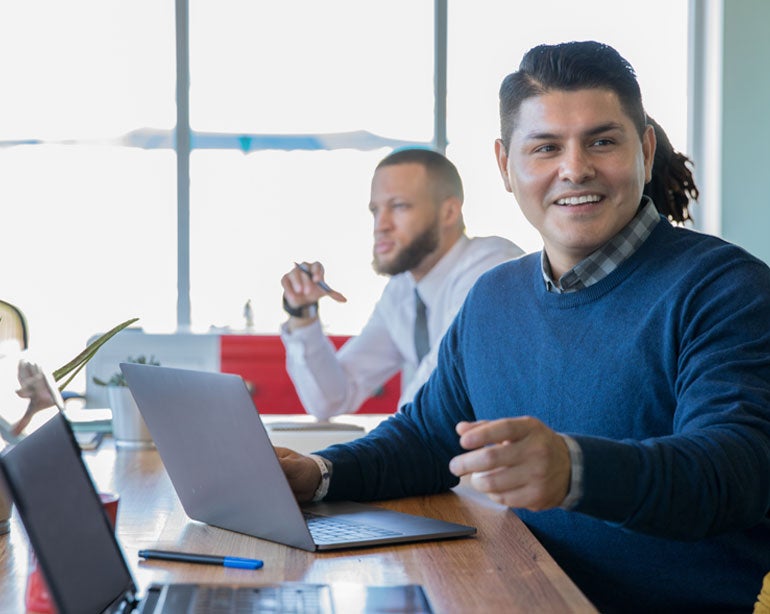 A Hispanic mail professional is setting in a meeting looking at his laptop and explaining material to another colleague.