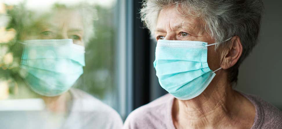 An older woman wears a surgical mask and is looking out the window while she is in quarantine because of COVID-19.