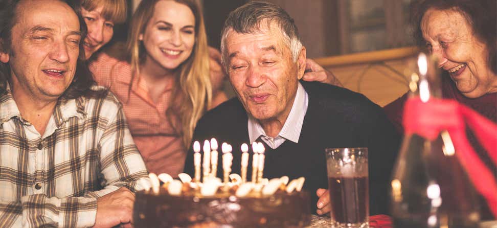 A senior man is sitting at the dining room table blowing out his candles for his 80th birthday with family.