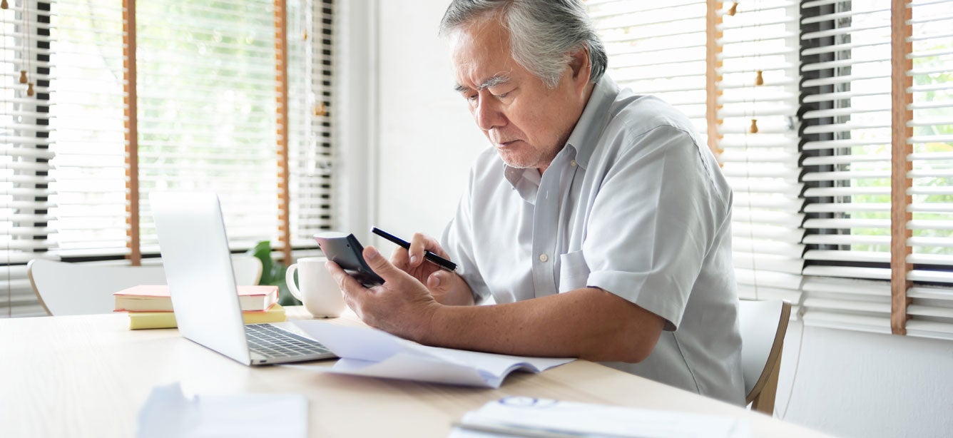 An older Asian man sits at his kitchen table, working on his finances in front of a computer and holding a calculator. 
