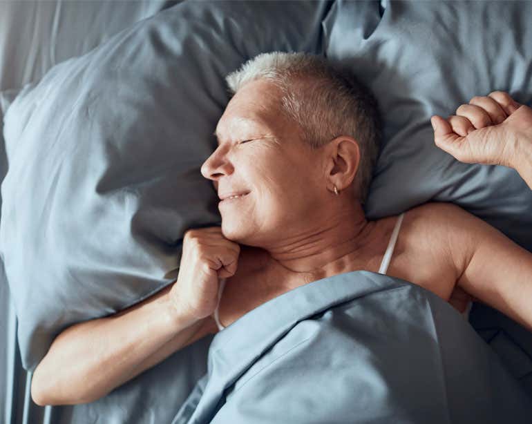 A senior woman is waking up after a restful night's sleep, smiling as the sun shines in her room.