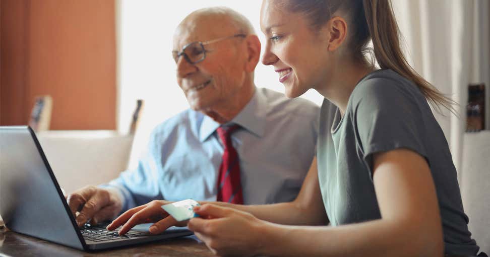 A younger benefits enrollment professional helps a senior find benefits options online. 