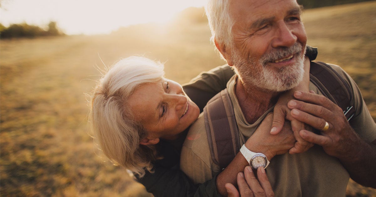A senior Caucasian couple is walking in a field at sunset, smiling and embracing each other. 