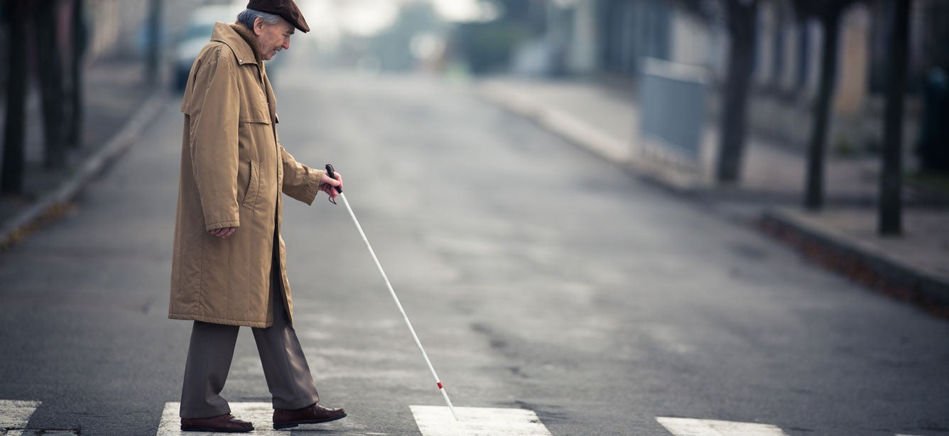 A senior man suffering from vision loss, wearing a khaki trench coat is walking across the street using walking stick.
