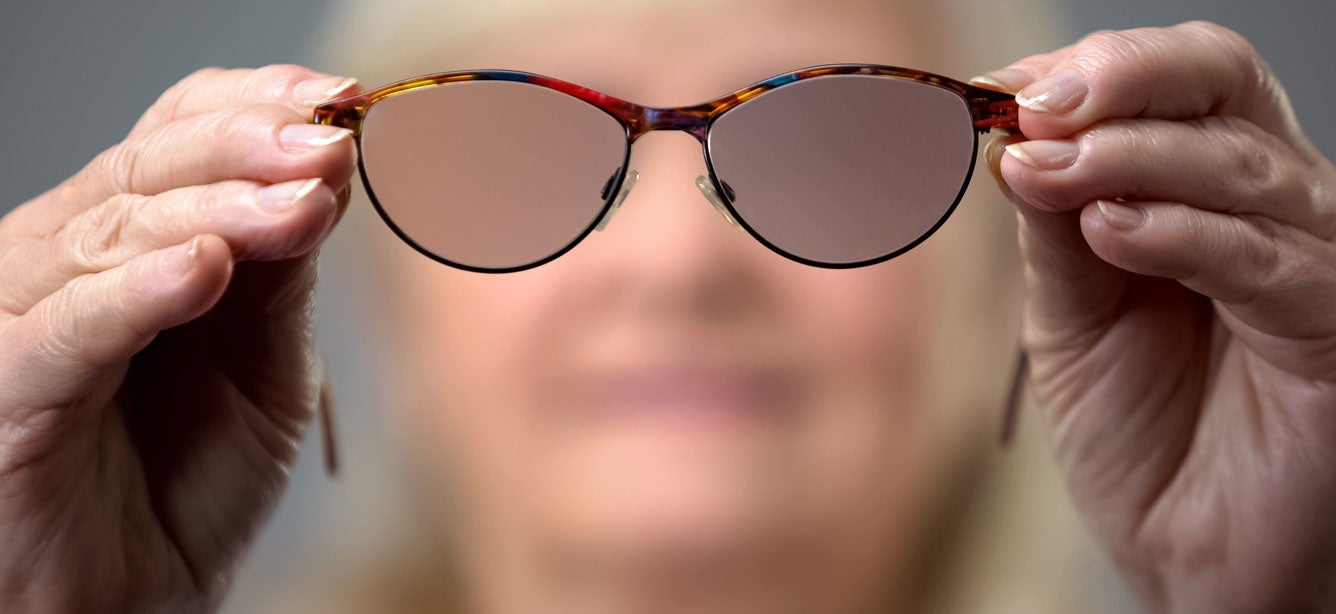 A senior Caucasian woman is holding blurred glasses up close to the camera, indicating the concept of vision loss.