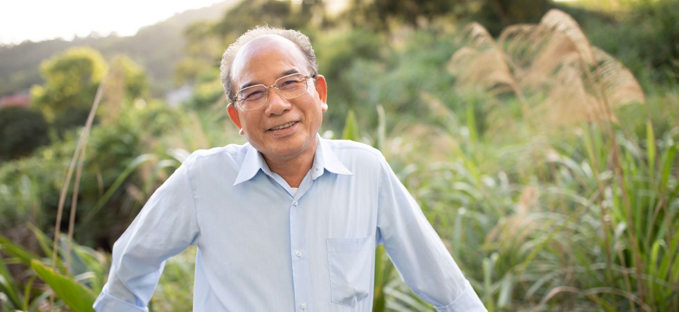An older Asian man smiles on the balcony of his house.