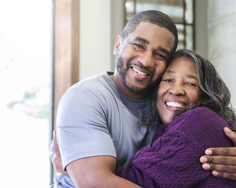 A younger Black man is hugging his older adult mother, both smiling and enjoying life.