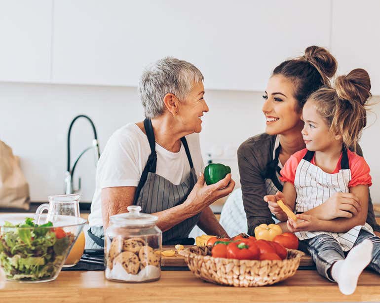 A senior woman is talking to her daughter and granddaughter at the island in her kitchen, smiling.