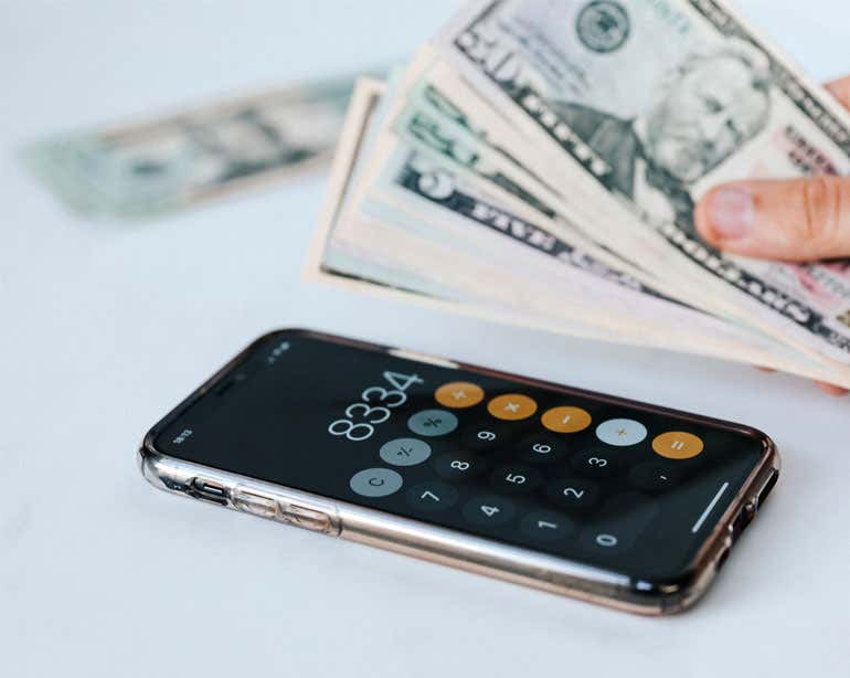 An older generation iPhone with a calculator on display sits next to hand holding a stack of dollar bills.