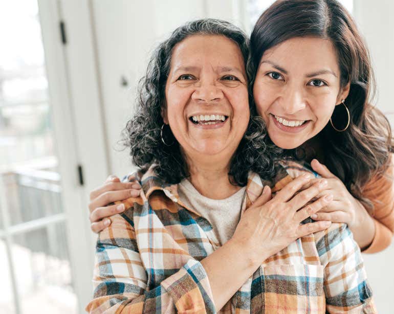 A younger caregiver hugs her senior Hispanic mother from behind, both are smiling.