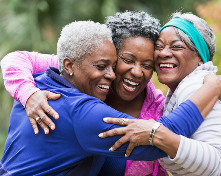 Three senior African American women are at a park hugging.