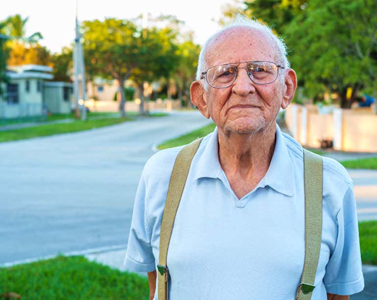 A senior man in a blue-collared shirt and suspenders stares straight on towards the camera with a depressed demeanor.