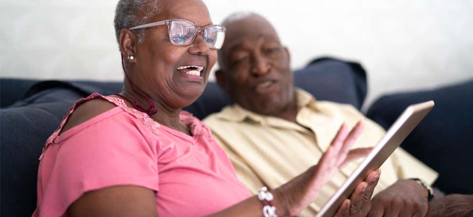 A Black senior woman and her husband are sitting on the couch looking at something on their tablet.