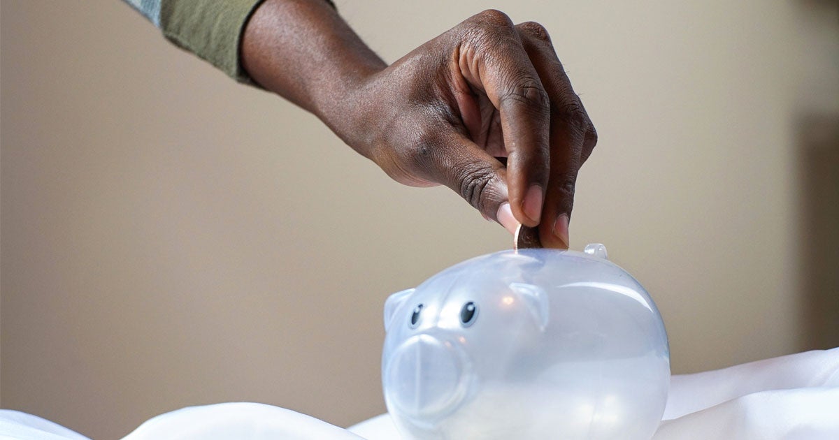 A Black senior hand is placing coins in a clear piggy bank.