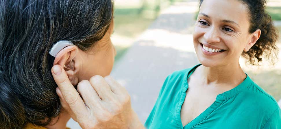A senior woman with black hair is adjusting her hearing aid while having a conversation with her younger daughter outside.