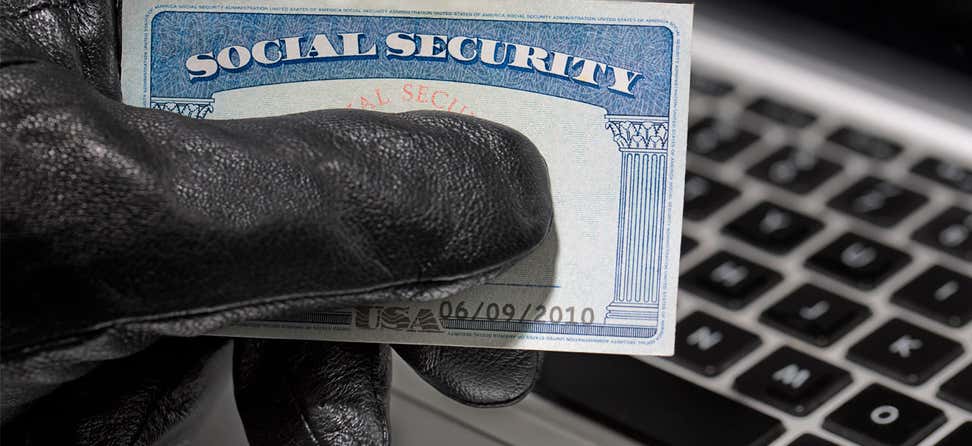 A thief with a black glove on holds a social security card while trying to access a stolen computer.