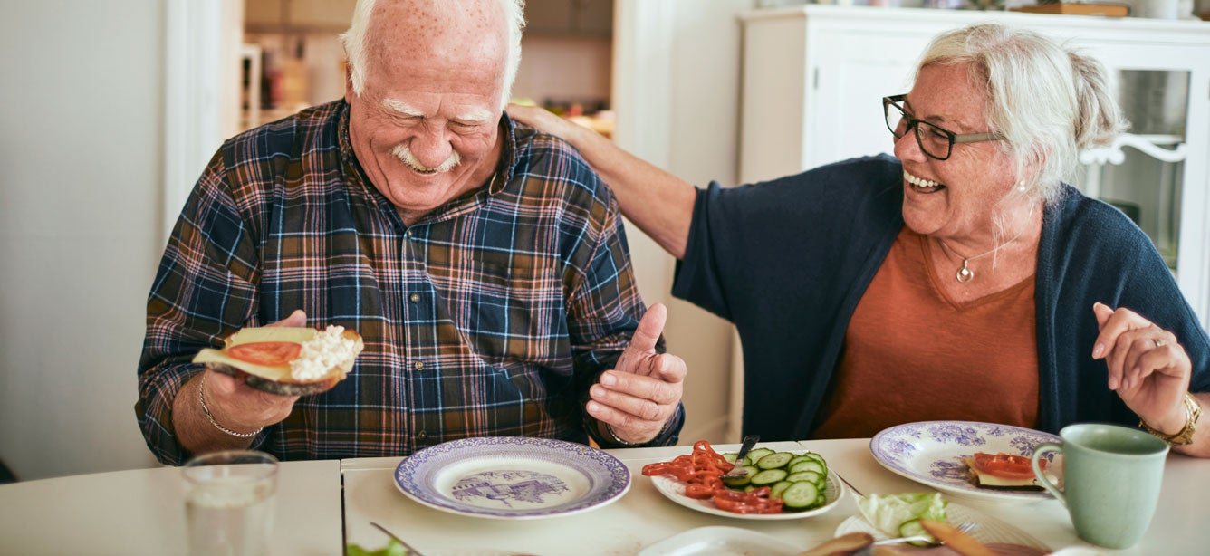 A senior couple is laughing at the dinner table while eating a healthy meal together.