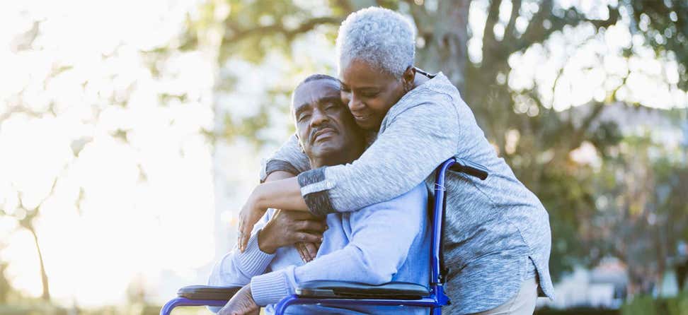 A Black senior woman embraces her husband who's sitting in a wheelchair. They're enjoying the beautiful day outside.