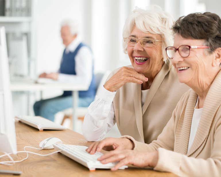 Two senior women are working at a computer together, smiling. There's an older man seen in the foreground also on a computer.