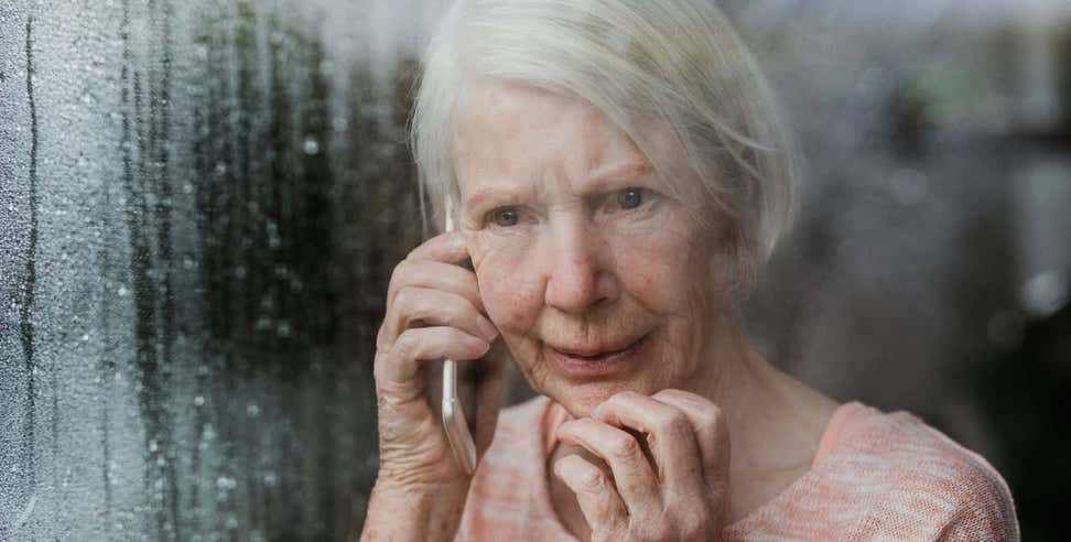 Older woman looking out window at storm while on cell phone with worried expression