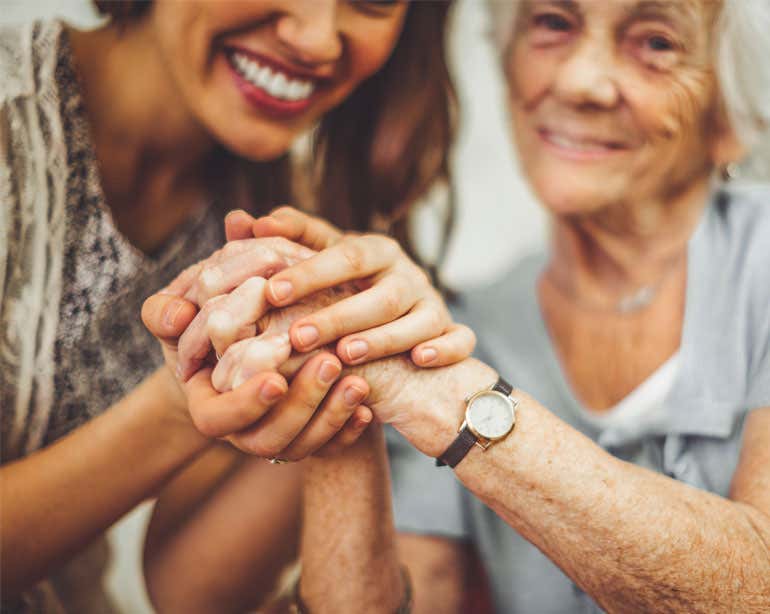 A younger female caregiver holds the hands of a senior Caucasian woman who is smiling.
