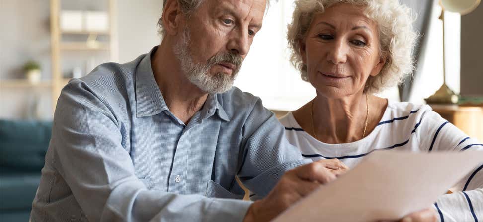 A senior man holds his medication record form, while seemingly confused, and discusses the content with his wife.