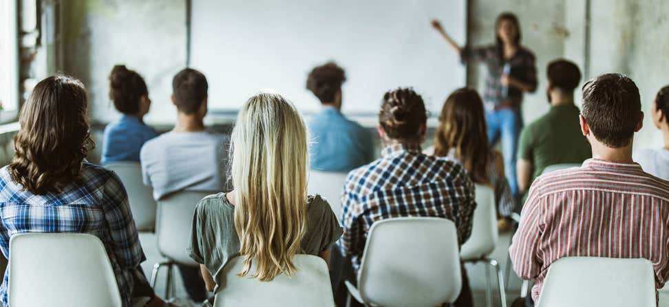 A group of professionals are sitting through a seminar while an instructor is up at the front giving a presentation.