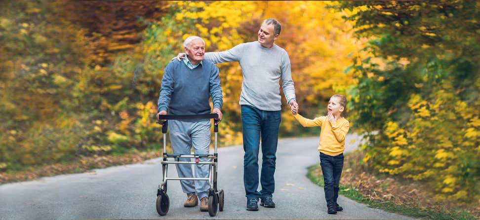 A middle aged man, along with his young son, is taking a stroll in the park with his elderly father that's using a walker.