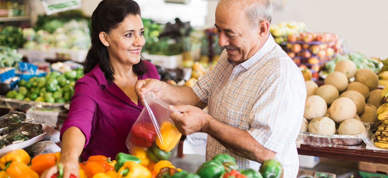 Older Hispanic couple is putting bell peppers into bag at grocery store together.