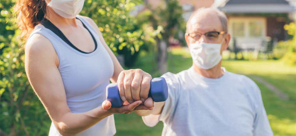 A senior man is getting exercise help from a caregiver outside, he's working on lifting a dumb bell weight for strength.