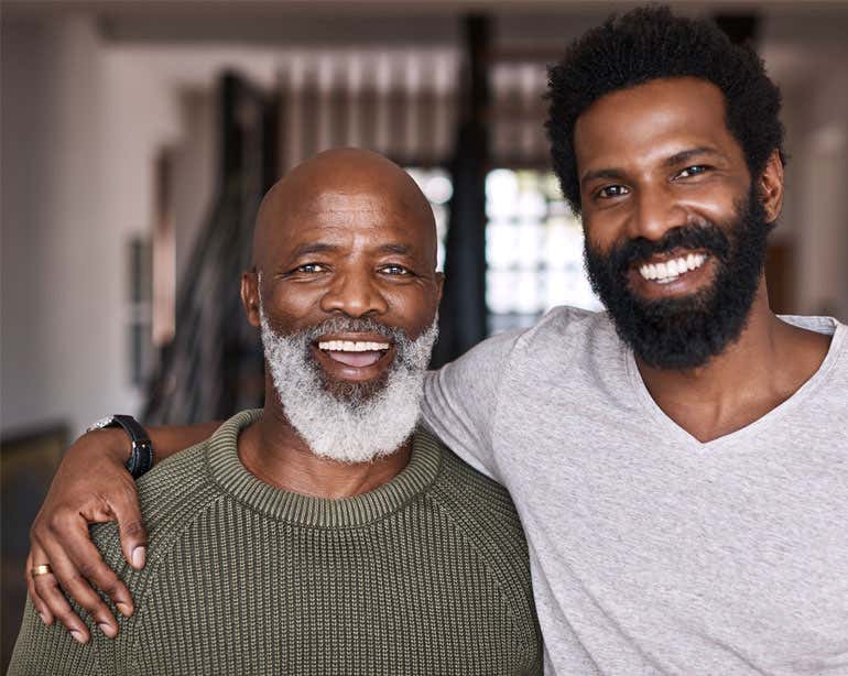 A Black younger caregiver is hugging his senior father, posing for the camera.