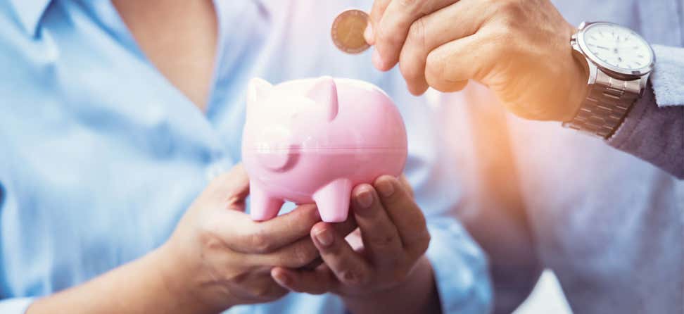 A senior Asian woman is holding a pink piggy bank while her husband gets ready to place a coin inside of it.