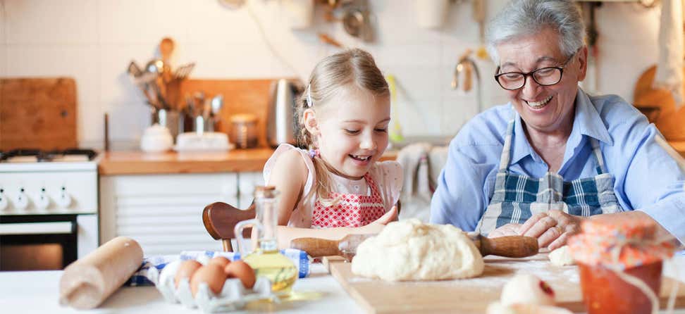 A grandmother wearing glasses is in her kitchen making dough with her granddaughter, both are smiling, having fun.