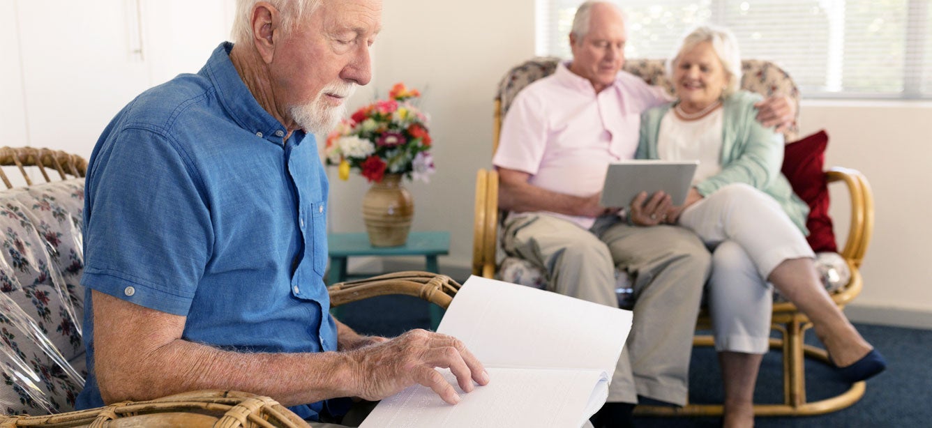 A Caucasian senior man is reading braille while sitting in a senior center with other older adults.